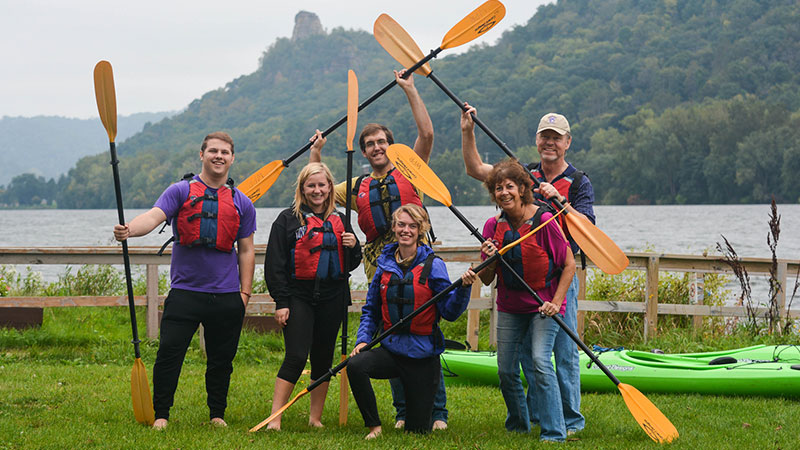 Outdoor photo a group of people with kayak paddles and a lake behind them